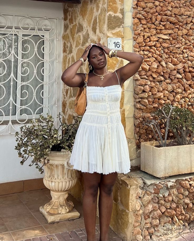 Mulher jovem de pele negra. Ela usa vestido branco de comprimento curto e botões na parte da frente.