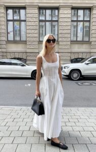 Mulher jovem, magra e de pele clara. Ela usa vestido branco longo, sapatilha preta e bolsa preta.
