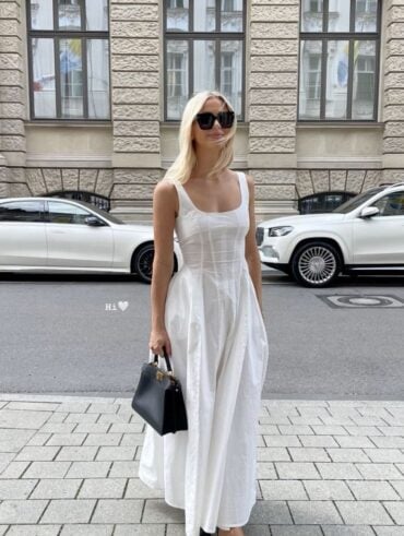 Mulher jovem, magra e de pele clara. Ela usa vestido branco longo, sapatilha preta e bolsa preta.