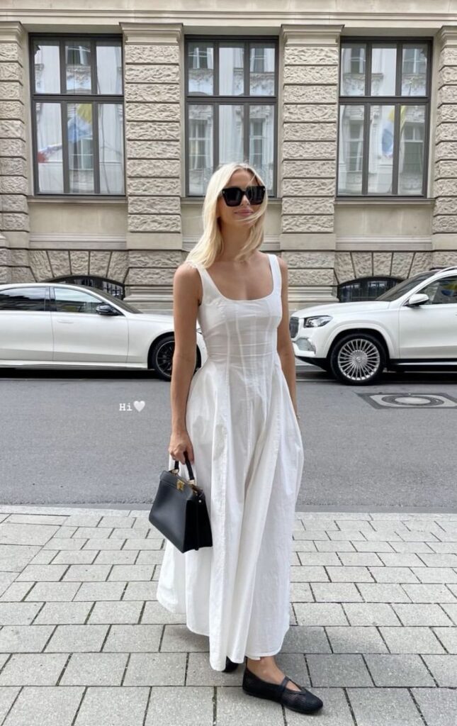 Mulher jovem, magra e de pele clara. Ela usa vestido branco longo, sapatilha preta e bolsa preta.
