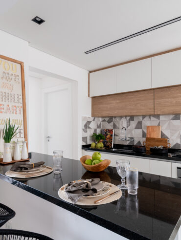 Cozinha com móveis brancos e em MDF que remete à madeira, com bancada preta, quadro e mesa posta