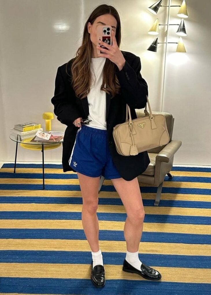 Mulher jovem, branca e magra, usando blazer preto com camiseta branca, short Adidas, mocassim com meia longa e segurando uma bolsa.
