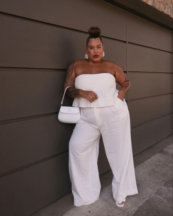 Mulher jovem, branca e plus size, usando cabelo preso em coque, blusa sem alça na cor branca, calça pantalona branca e uma bolsa na mesma tonalidade, com brincos grandes.