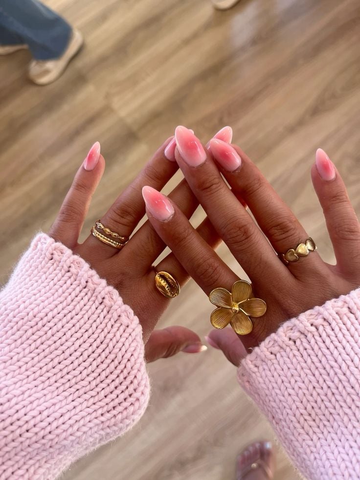 Mão de pele clara-média, com anéis em dourado e unha amendoada rosa aura 