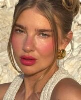Mulher de pele clara, com regata branca, cabelo meio preso, acessórios dourados e blush rosa