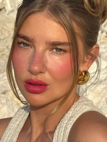 Mulher de pele clara, com regata branca, cabelo meio preso, acessórios dourados e blush rosa