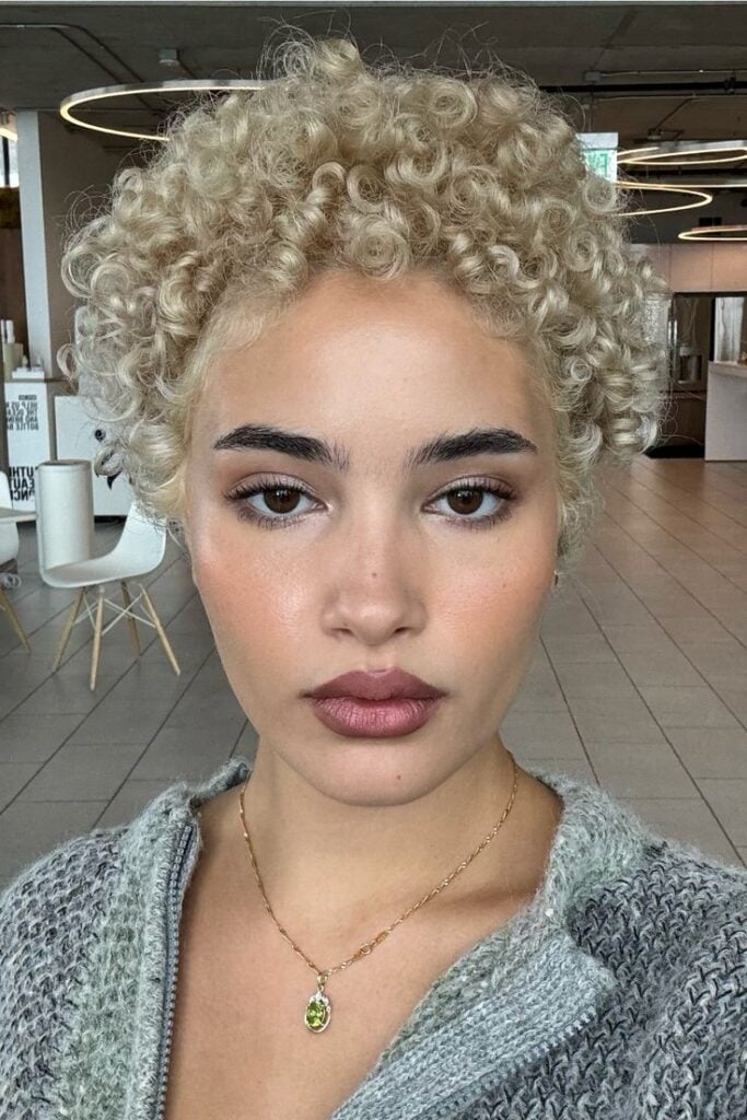 Mulher de pele clara, com agasalho cinza, colar dourado e cabelo pixie loiro e cacheado