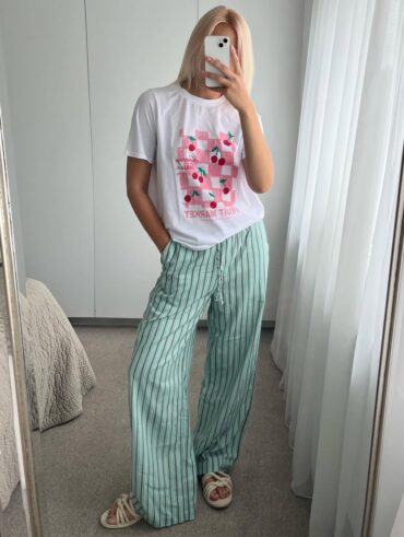 Mulher magra e de pele clara, com cabelo loiro solta, blusa estampada, calça de linho listrada e calçado off-white