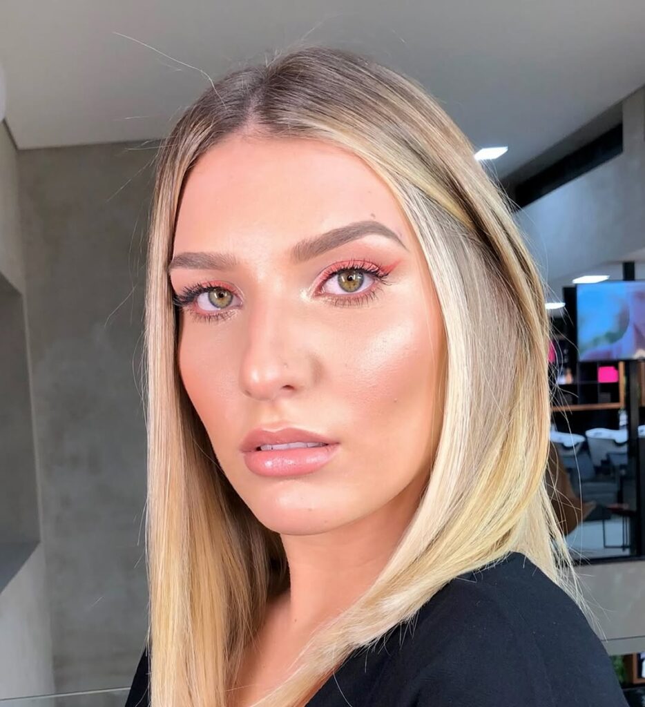 Mulher de pele branca, com roupa preta, cabelo loiro e delineado coral