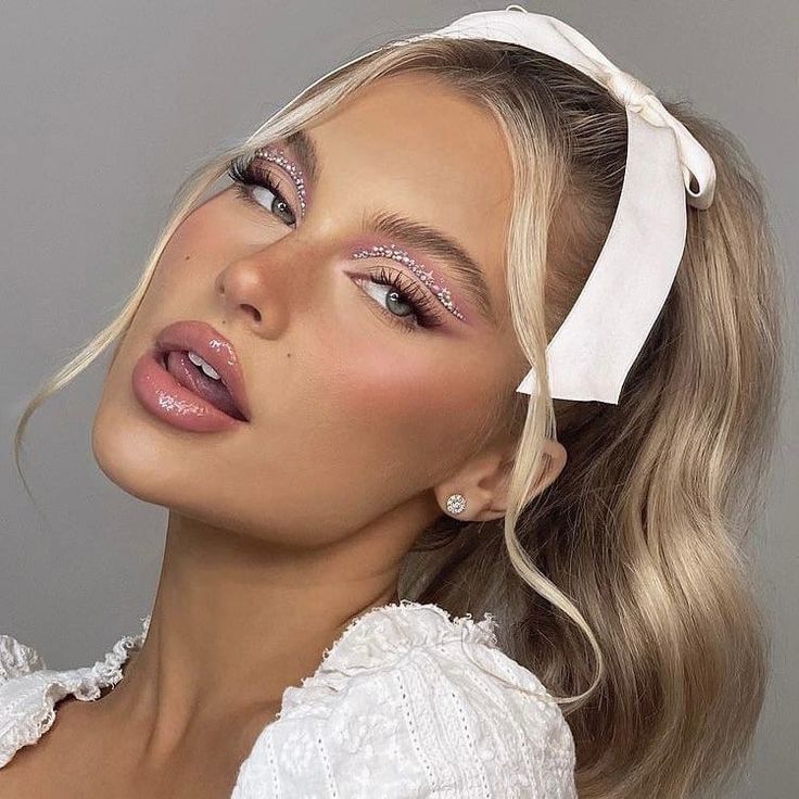 Mulher de pele branca, com vestido branco, cabelo loiro meio-preso com laço e maquiagem com tons de rosa e delineado de pedrinha 