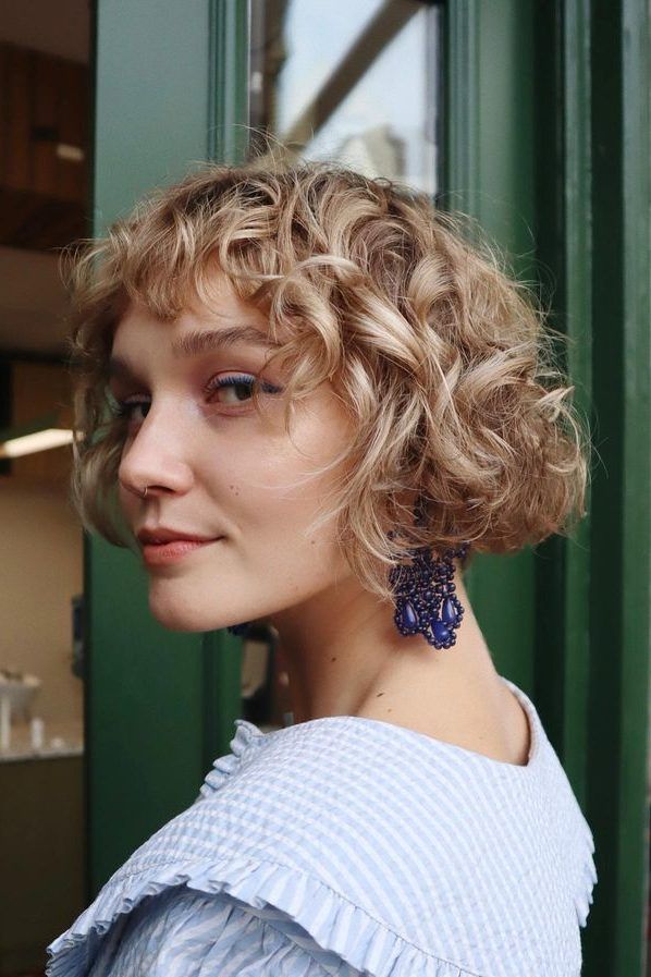 Mulher de pele clara, com roupa azul-claro e cabelo loiro e cacheado com corte french bob