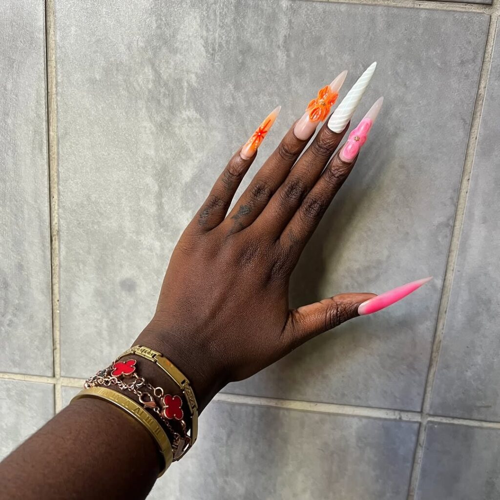 Mão de pele preta, com acessórios coloridos e em dourado e unha stiletto colorida e com itens em 3D
