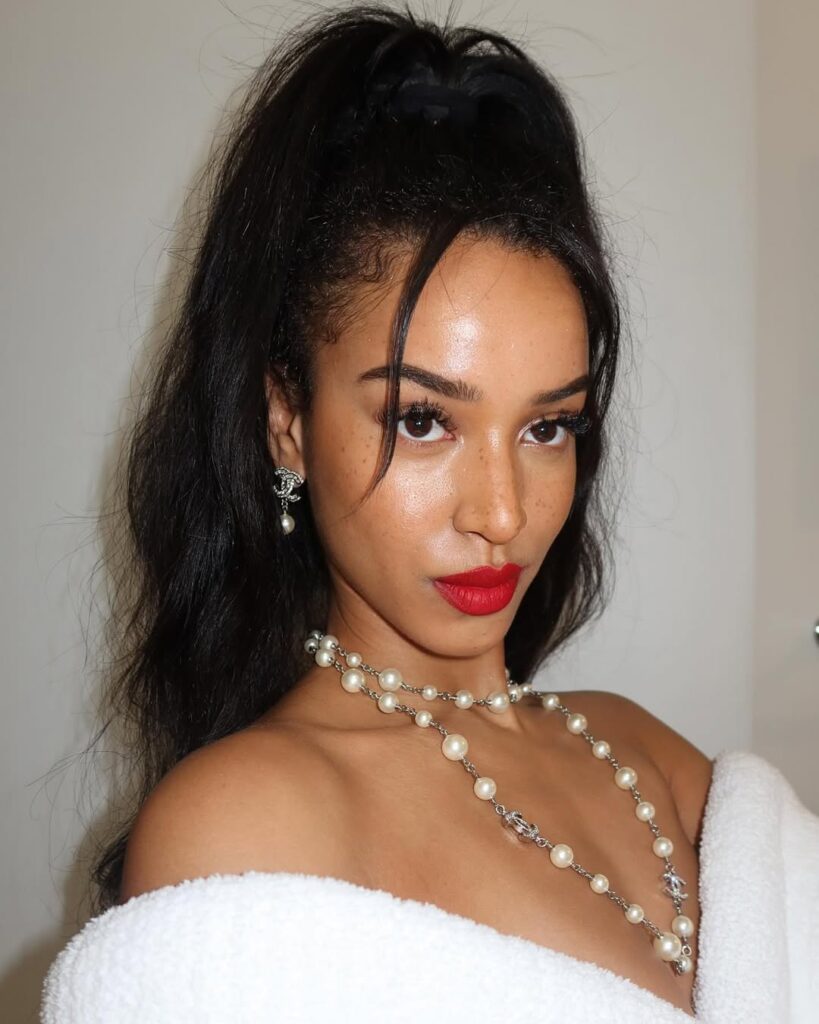 Mulher de pele preta, com cabelo escuro meio-preso, colar de pérolas e maquiagem com boca vermelha