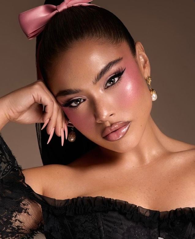 Mulher de pele clara, com cabelo escuro preto com laço rosa, roupa preta e maquiagem com bochechas rosa e lábios contornados 