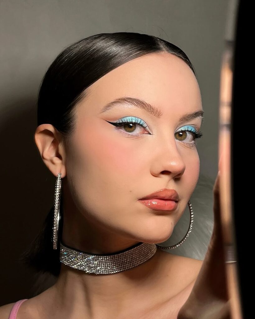 Mão de pele clara, com cabelo preso, acessórios brilhosos e maquiagem com sombra azul
