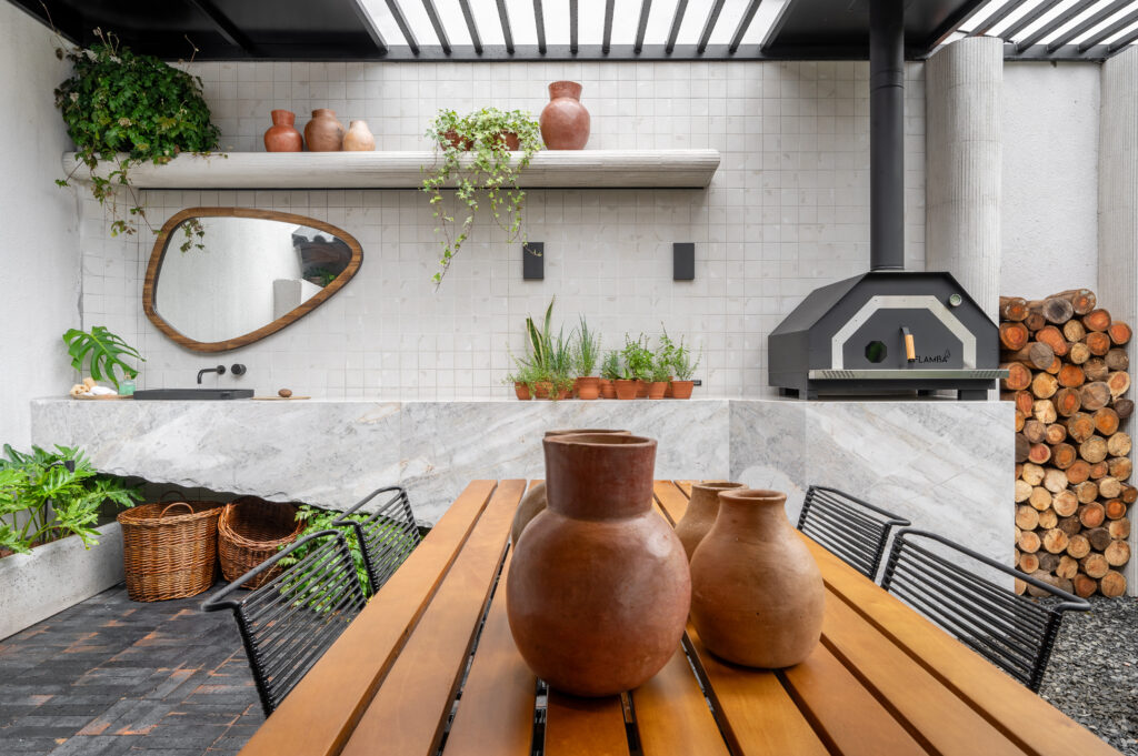 Ambiente externo com mesa de madeira, cadeiras em metal preto, vasos de barro e forno a lenha