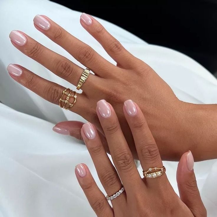 Mãos de pele clara, com anéis em dourado e unha curta e quadrada rosa perolada