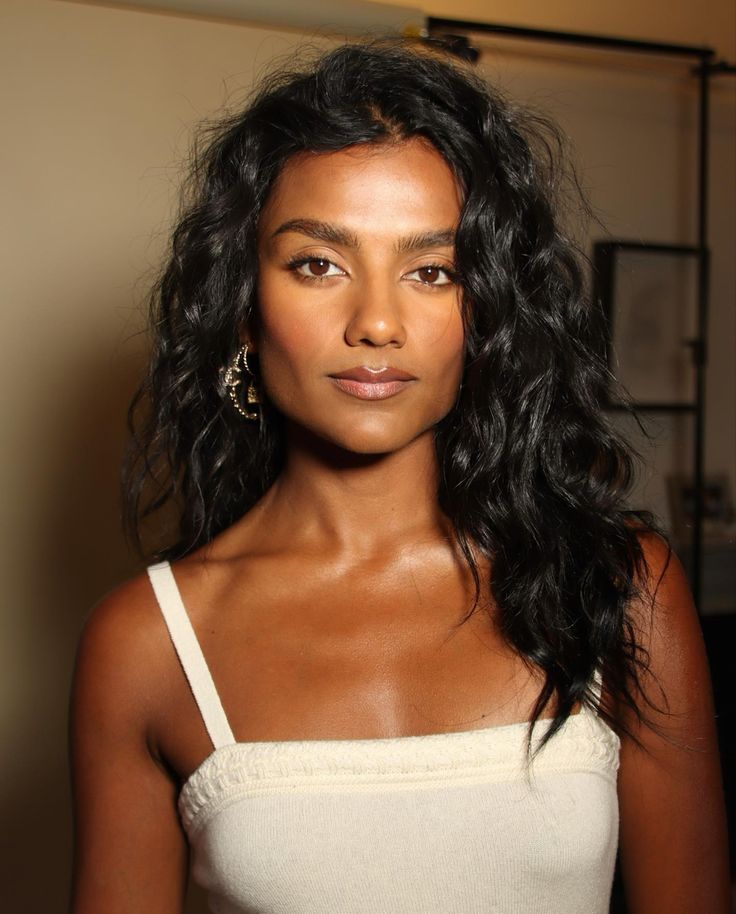 Simone Ashley com blusa branca, acessórios em dourado e cabelo escuro cacheado