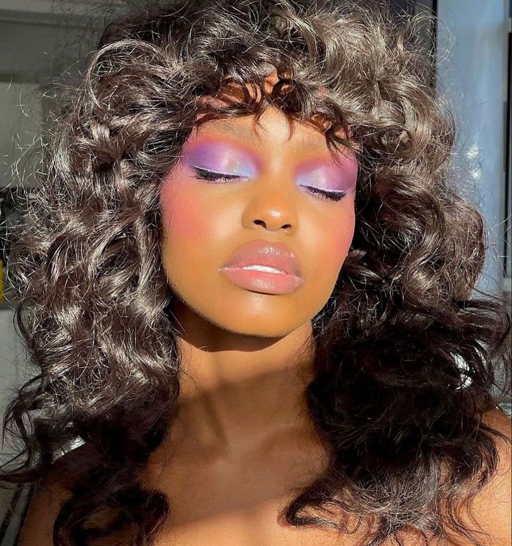 Mulher de pele preta, com cabelo cacheado escuro com franja e maquiagem com sombra colorida