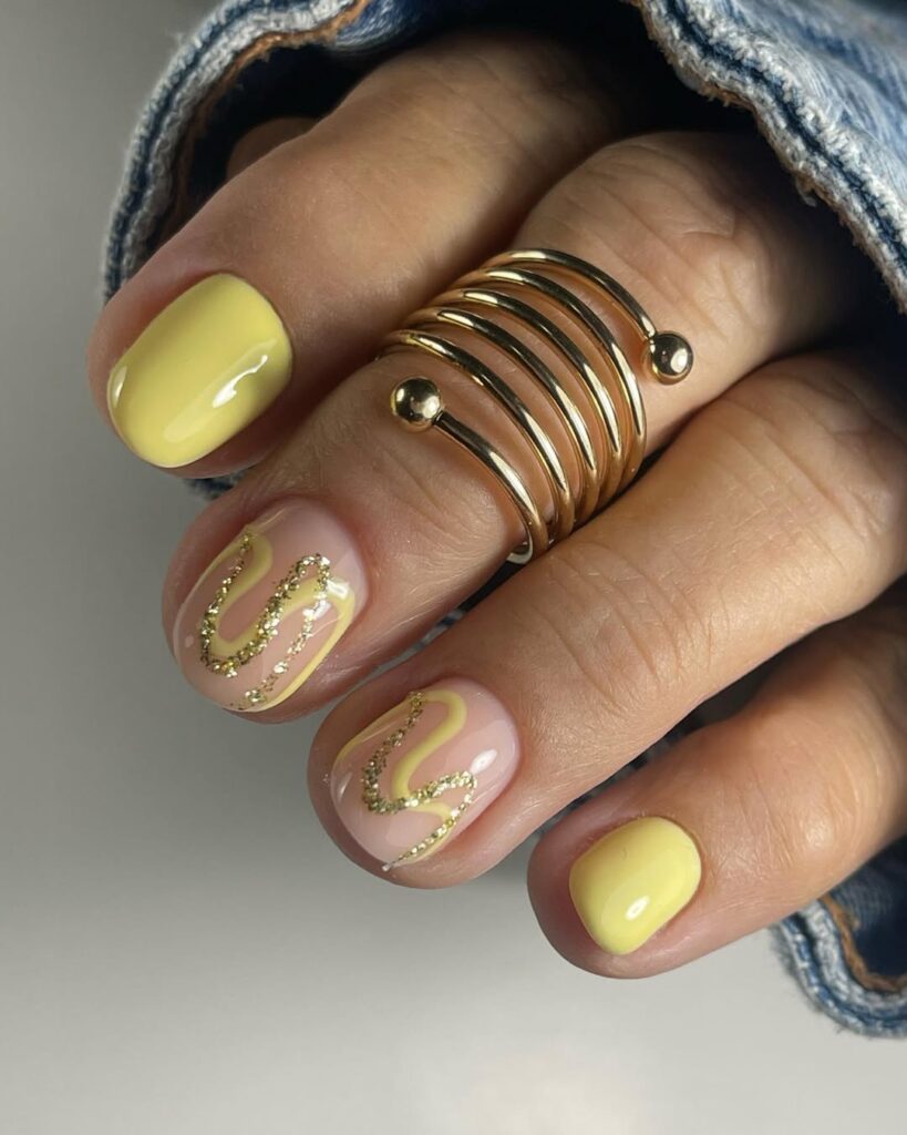 Mão de pele clara, com acessório em dourado e unha curta e redonda amarelo-manteiga com linhas fluidas com essa cor e glitter