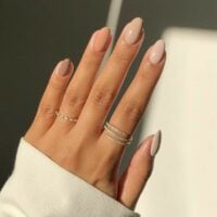 Mão de pele clara, com anéis em dourado e unha amendoada bege degradê