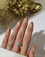 Mão de pele clara, com anéis brilhantes e unha amendoada nude com pontas com glitter dourado e estrela
