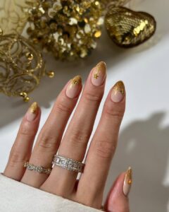 Mão de pele clara, com anéis brilhantes e unha amendoada nude com pontas com glitter dourado e estrela