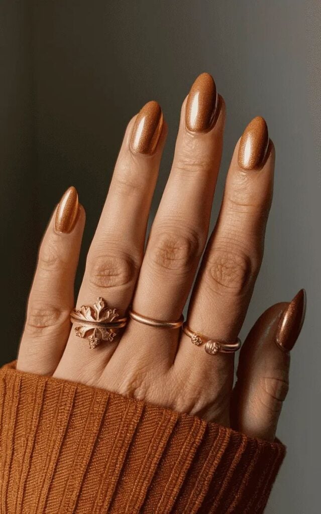 Mão de pele clara, com acessórios em dourado e unha amendoada terracota com brilho