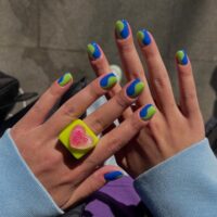 Mãos de pele clara, com anel colorido, unha curta com esmalte verde e azul