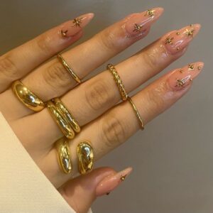 Mão de pele clara, com anéis em dourado e unha amendoada nude com charms em dourado