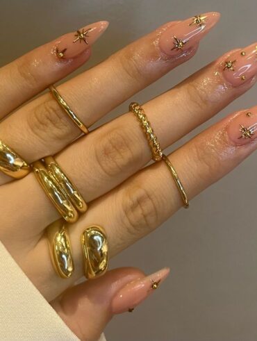 Mão de pele clara, com anéis em dourado e unha amendoada nude com charms em dourado