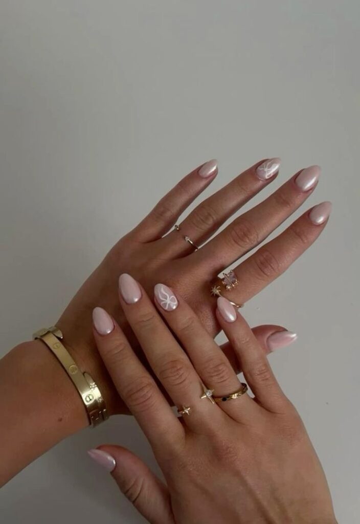 Mãos de pele clara, com acessórios em dourado e unha amendoada glazed com laço branco 