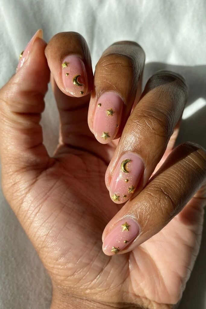 Mão de pele preta, com unha curta e amendoada nude com charms em dourado