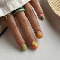 Mão de pele clara, com anel dourado e colorido e unhas curtinhas com 'estampa' de fruta