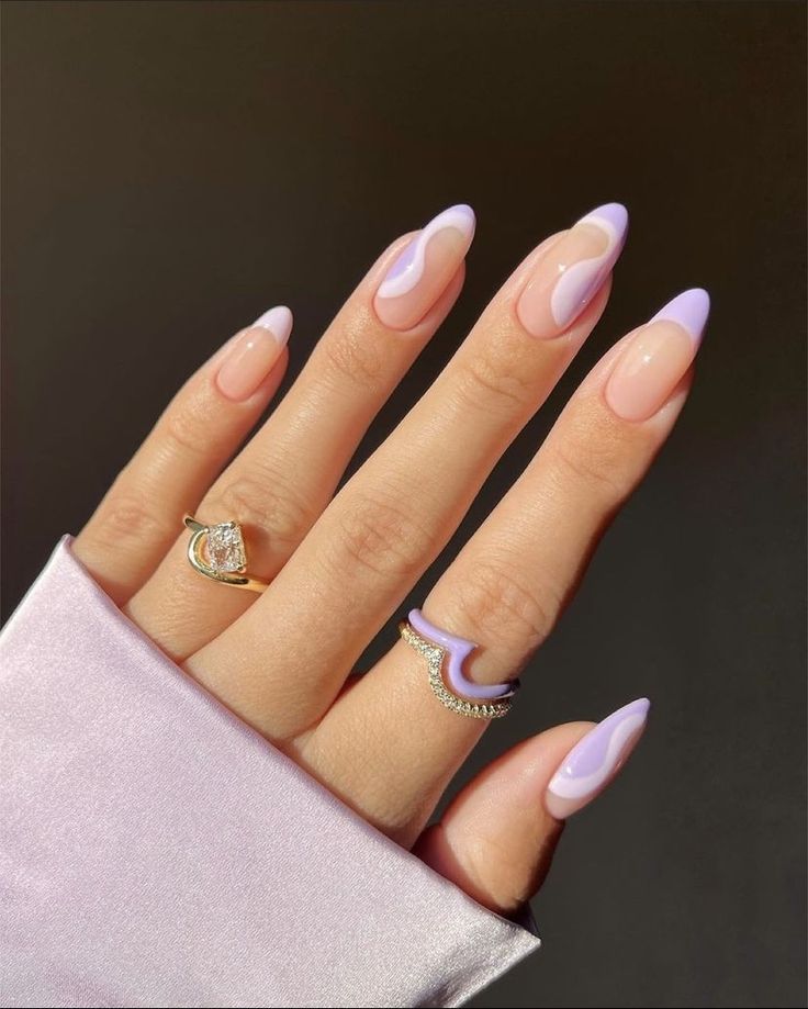 Mão de pele clara, com anéis em dourado e coloridos, e unha amendoada com esmalte lilás e branco 