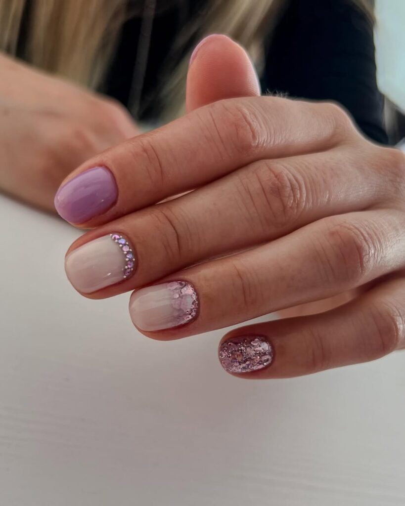 Mão de pele clara, com unha curta e redonda lilás, com glitter e strass