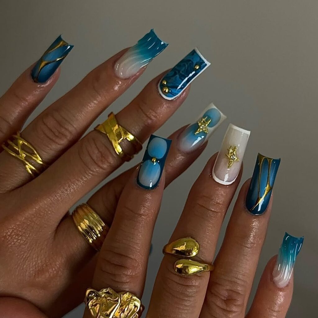 Mãos de pele clara, com anéis em dourado e unha quadrada azul com branco, detalhes em dourado e em alto relevo 