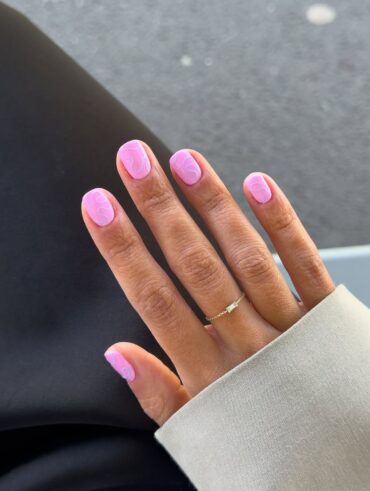 Mão de pele clara, com anel fininho e unha quadrada rosa com linhas fluidas coloridas