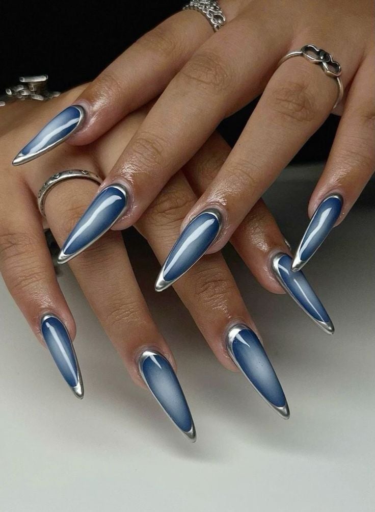 Mãos de pele clara-média, com acessórios em prata e unha stiletto aura azul com "moldura" em prata 