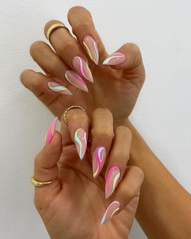 Mão de pele clara, com anéis em dourado e unha stiletto nude com linhas fluidas em tons pastel 