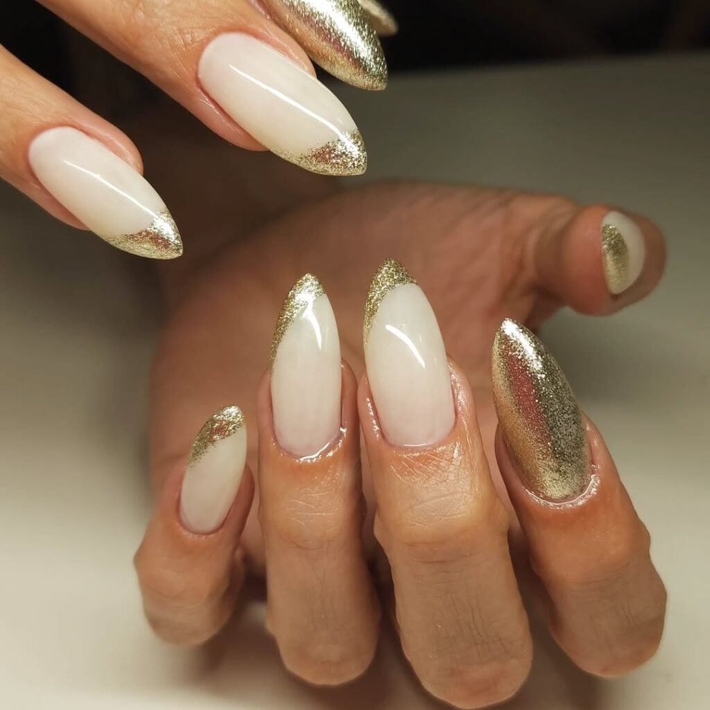 Mãos de pele clara, com unha stiletto com francesinha dourada com glitter e unha só com glitter