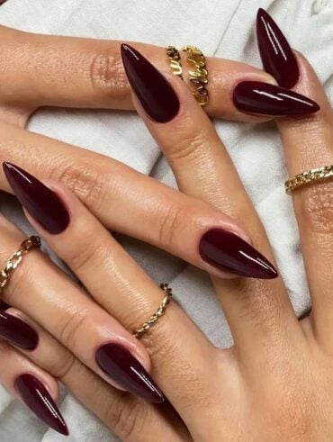 Mãos de pele clara, com anéis em dourado e unha stiletto marrom avermelhado