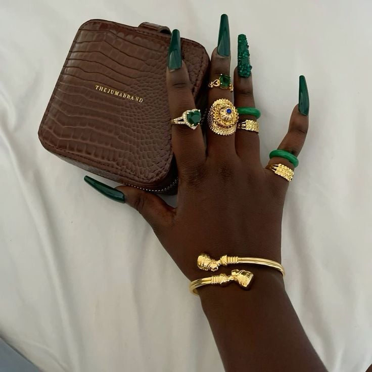 Mão de pele preta, com acessórios em dourado e unha unha quadrada verde-esmeralda
