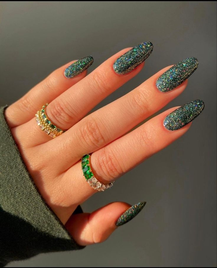 Mão de pele clara, com anéis coloridos e unha amendoada verde-esmeralda com glitter