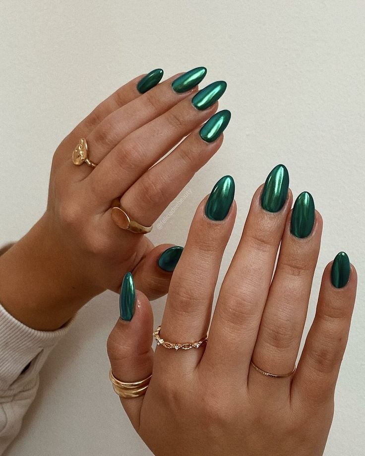 Mãos de pele clara, com anéis em dourado e unha amendoada verde-esmeralda cromada