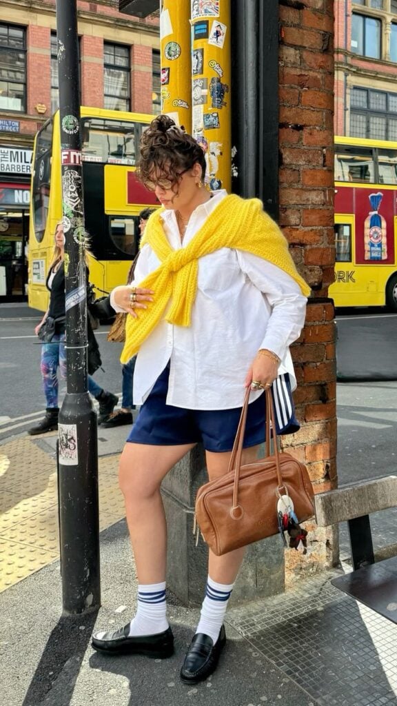 Mulher midi size e branca, usando cabelo preso, camisa branca com cardigan amarelo nos ombros, short Adidas, segurando uma bolsa marrom, meia longa e mocassim.