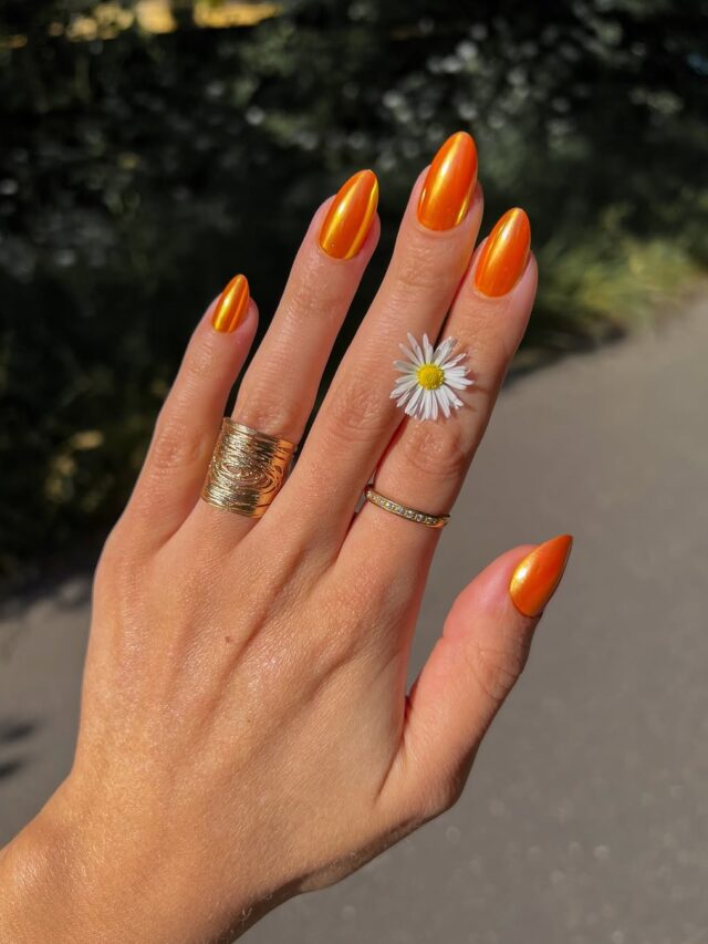 Mão de pele clara, com anéis dourados e unha amendoada laranja metalizada