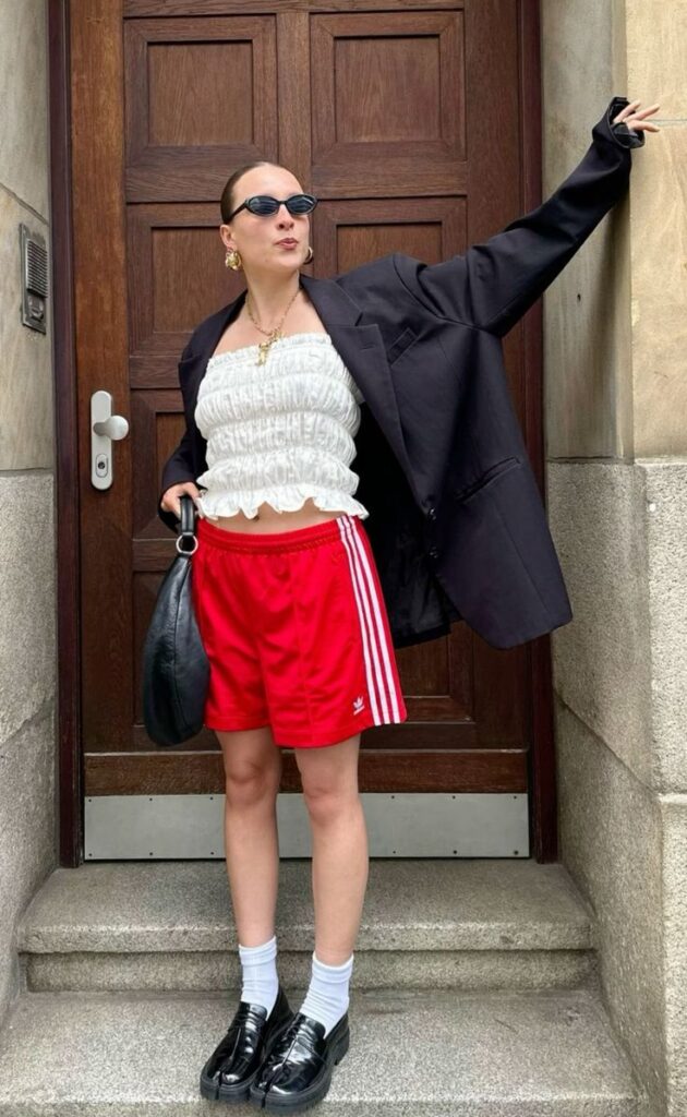 Mulher jovem, branca e magra, usando óculos de sol, blazer preto, blusa branca, short Adidas e mocassim.
