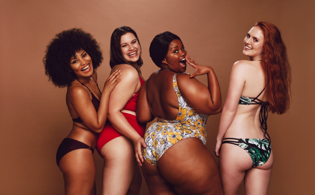 Grupo de mulheres de diferentes raças, tipos físicos e tamanhos em trajes de banho, juntas e rindo contra um fundo cinza. Mulheres diversas em biquínis olhando para a câmera.