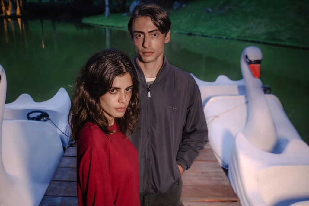 Julia Dalavia e Jaffar Bambirra em uma ponte, com pedalhinhos ao fundo